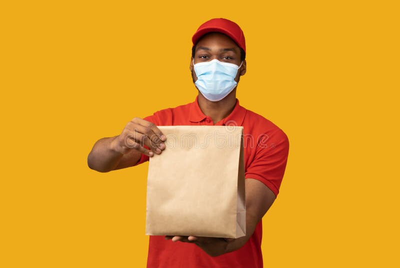 Food Delivery. Black courier in medical face mask and red cap holding and giving craft paper package from restaurant with products, isolated on yellow background. Shipment service during pandemic. Food Delivery. Black courier in medical face mask and red cap holding and giving craft paper package from restaurant with products, isolated on yellow background. Shipment service during pandemic