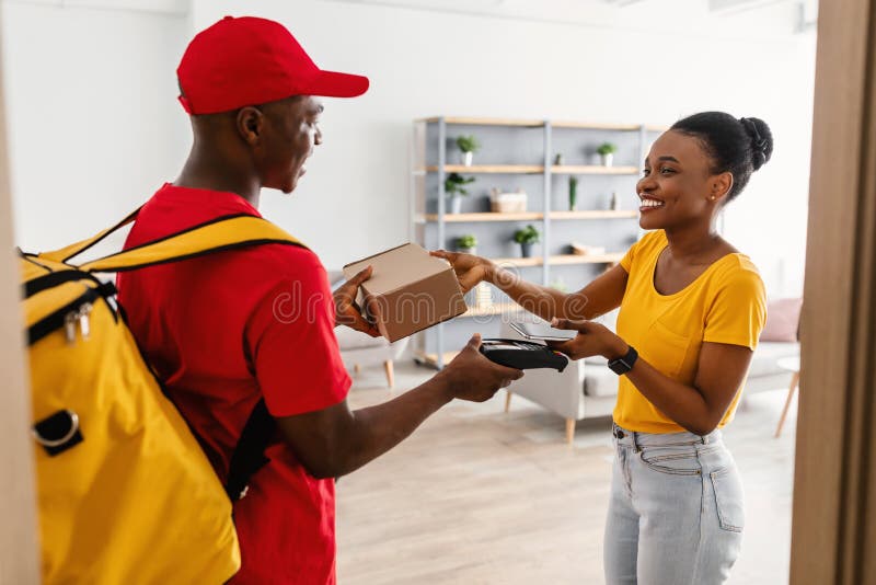 Door Delivery. Happy Black Customer Lady Receiving And Paying For Package Box With Phone Taking Parcel From Courier Guy In Red Uniform Standing In Doorway At Home. Modern Shopping Concept. Door Delivery. Happy Black Customer Lady Receiving And Paying For Package Box With Phone Taking Parcel From Courier Guy In Red Uniform Standing In Doorway At Home. Modern Shopping Concept