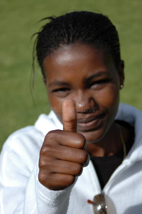 A black African American young woman head portrait with smiling expression in her pretty face showing her brown hand with the thumb up (focus on thumb) outdoors. A black African American young woman head portrait with smiling expression in her pretty face showing her brown hand with the thumb up (focus on thumb) outdoors