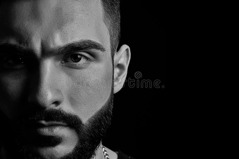 Black-and-white close-up of a dramatic portrait of a young serious guy, a musician, singer, rapper with a beard in black clothes . half a face on a black isolated background. Black-and-white close-up of a dramatic portrait of a young serious guy, a musician, singer, rapper with a beard in black clothes . half a face on a black isolated background.