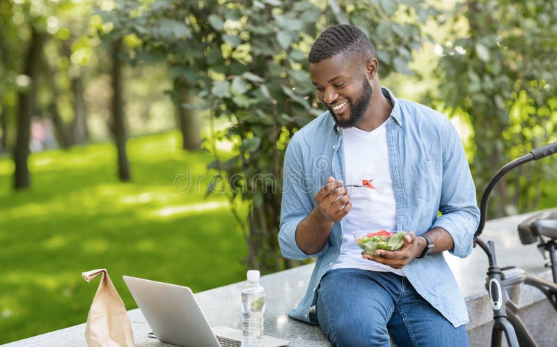 Lunch Fun. Young Black Guy Eating Takeaway Salad Outdoors And Watching Videos On Laptop, Relaxing On Bench In Park. Lunch Fun. Young Black Guy Eating Takeaway Salad Outdoors And Watching Videos On Laptop, Relaxing On Bench In Park