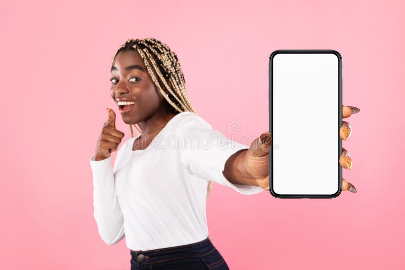 Portrait of cheerful young african american woman holding mobile phone with white blank screen in hand, showing device. Gadget with empty free space for mock up, selective focus, blurred background. Portrait of cheerful young african american woman holding mobile phone with white blank screen in hand, showing device. Gadget with empty free space for mock up, selective focus, blurred background