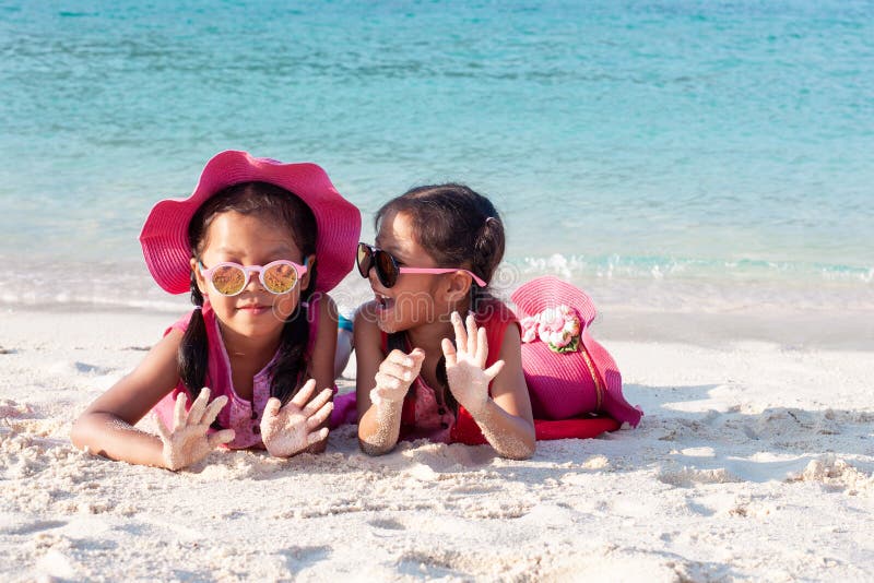 Zwei asiatische Kindermädchen, die rosa Hut und die Sonnenbrille zusammen spielt mit Sand auf dem Strand nahe dem schönen Meer stockfotografie