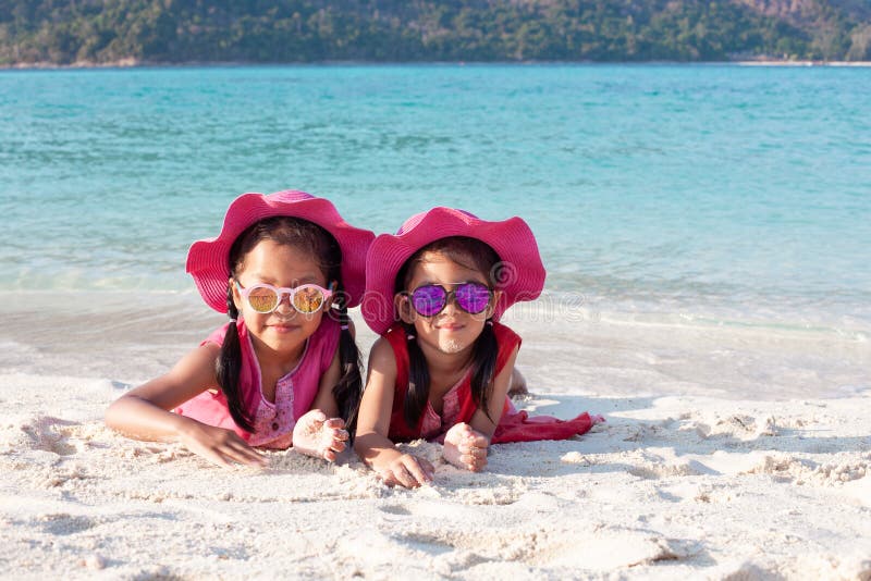 Zwei asiatische Kindermädchen, die rosa Hut und die Sonnenbrille zusammen spielt mit Sand auf dem Strand nahe dem schönen Meer stockfoto