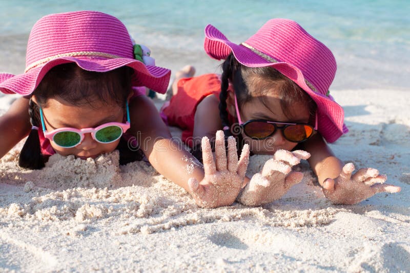 Zwei asiatische Kindermädchen, die rosa Hut und die Sonnenbrille zusammen spielt mit Sand auf dem Strand nahe dem schönen Meer lizenzfreie stockbilder