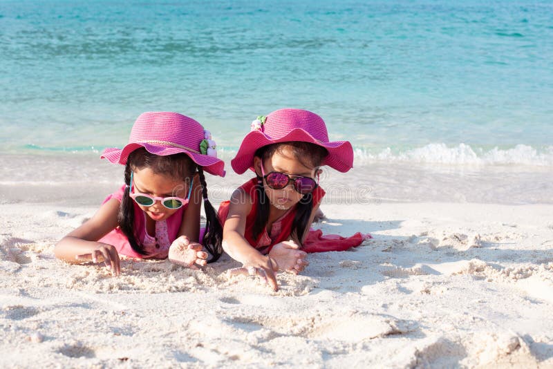 Zwei asiatische Kindermädchen, die rosa Hut und die Sonnenbrille zusammen spielt mit Sand auf dem Strand nahe dem schönen Meer stockfotos