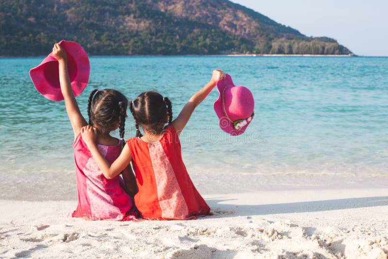 Zwei asiatische kleines Kindermädchen, die den Hut zusammen sitzt und spielt auf dem Strand nahe dem schönen Meer im Sommer hal lizenzfreies stockfoto