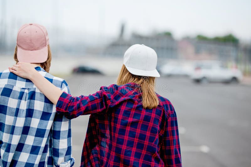 Zwei blonde Mädchen, die karierte Hemden, Kappen und Denimkurze hosen tragen, stehen mit ihren Rückseiten auf dem leeren Parkplat stockbilder Zwei blonde Mädchen, die karierte Hemden, Kappen und Denimkurze hosen tragen, stehen mit ihren Rückseiten auf dem leeren Parkplat stockbilder
