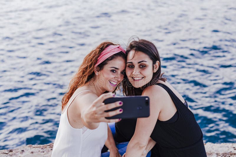 Zwei Freundinnen, die durch den Strand unter Verwendung des Handys, ein selfie und ein Lächeln nehmend sitzen Technologie- und Le stockfotografie Zwei Freundinnen, die durch den Strand unter Verwendung des Handys, ein selfie und ein Lächeln nehmend sitzen Technologie- und Le stockfotografie