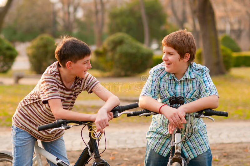 Two happy teenage boys talk in the park while resting on their bicycles. Two happy teenage boys talk in the park while resting on their bicycles.