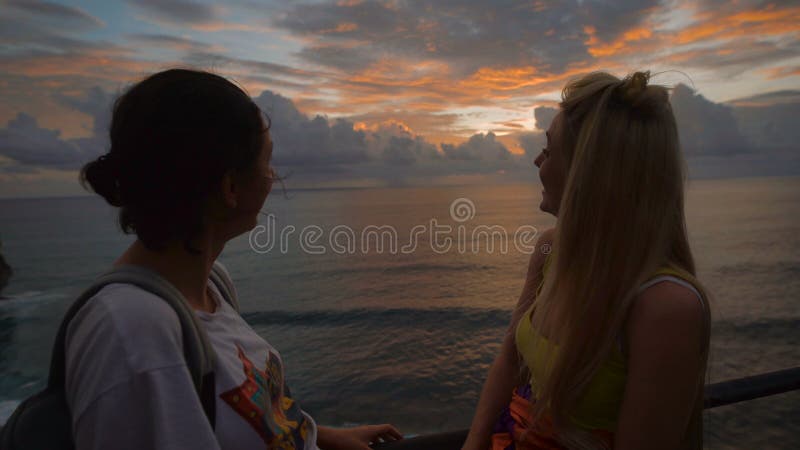 Zwei glückliche weibliche Reisende, die bei dem Sonnenuntergang gegen einen Hintergrund des Meeres sprechen und lachen Blondine u