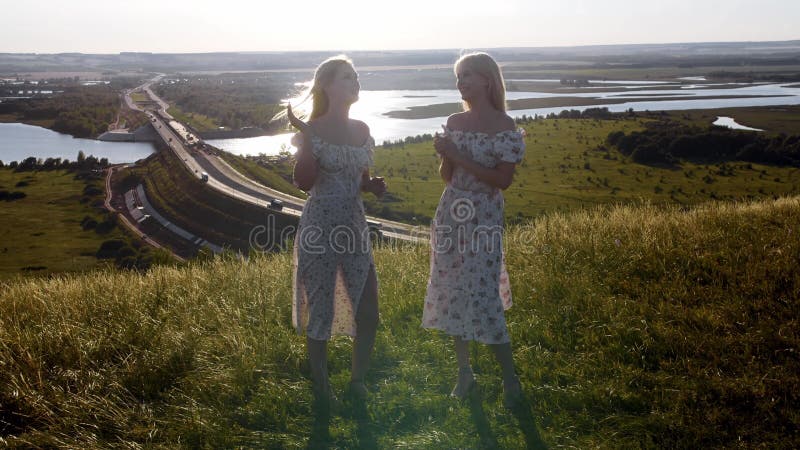 Zwei junge Blondinen in hellen Sommerkleiden, die auf einem Hügel sprechen