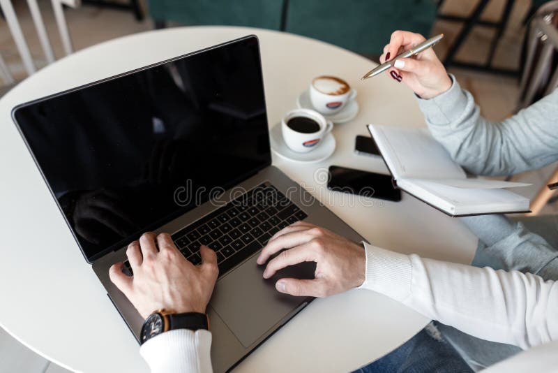 Zwei junge Geschäftsleute sitzen an einem Tisch in einem modernen Büro mit Kaffee und einem Laptop mit einem Notizbuch und einem stockfotografie Zwei junge Geschäftsleute sitzen an einem Tisch in einem modernen Büro mit Kaffee und einem Laptop mit einem Notizbuch und einem stockfotografie