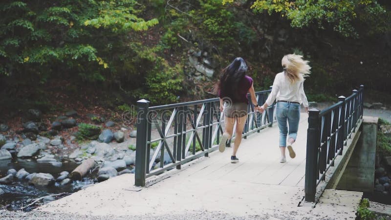 Zwei Mädchen halten Hände und Volkslauf über der Brücke über einem sauberen schnellen Fluss, einer Blondine und einem Brunette mi