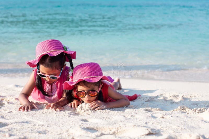 Zwei nette asiatische Kindermädchen, die rosa Hut und die Sonnenbrille zusammen spielt mit Sand auf dem Strand nahe dem schönen lizenzfreie stockfotografie
