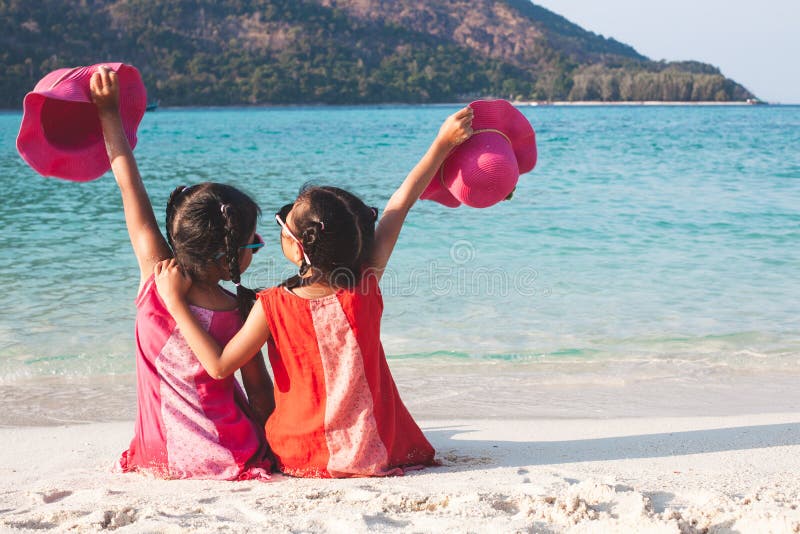 Zwei nette asiatische kleines Kindermädchen, die den Hut zusammen sitzt und spielt auf dem Strand nahe dem schönen Meer halten lizenzfreie stockfotos