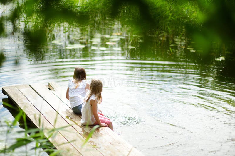 Zwei nette kleine Mädchen, die auf einer hölzernen Plattform durch den Fluss oder den See eintauchen ihre Füße im Wasser am warmen Sommertag sitzen Familientätigkeiten im Sommer