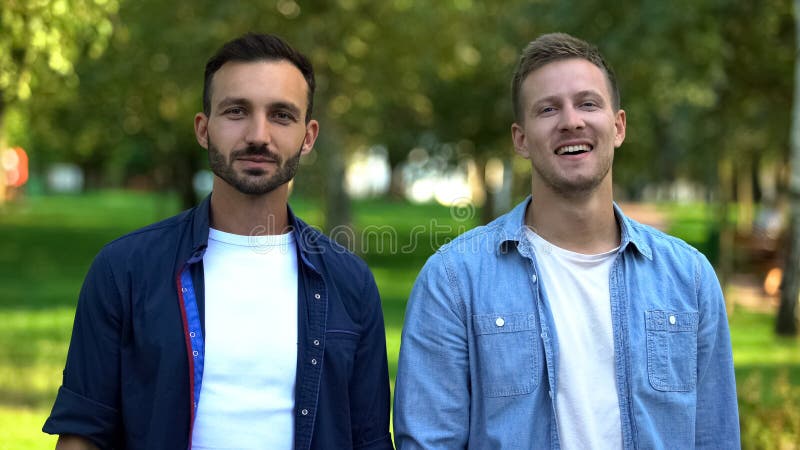 Zwei positive junge Männer, die eine Kamera sehen, männliche Freunde, die zusammen im Park sitzen stockfotos Zwei positive junge Männer, die eine Kamera sehen, männliche Freunde, die zusammen im Park sitzen stockfotos