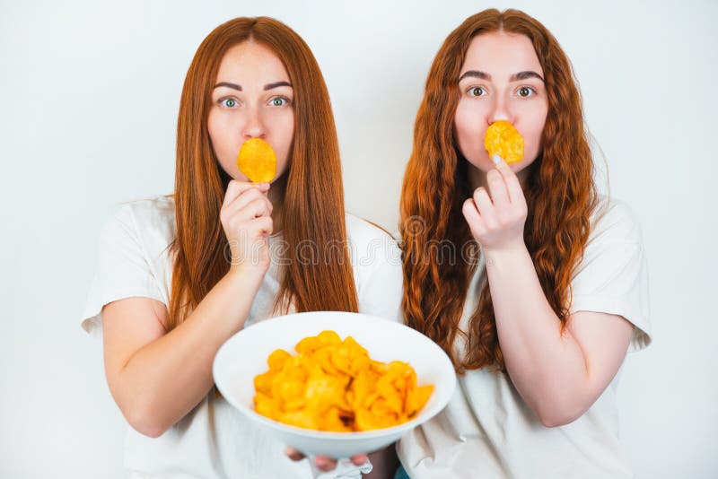 Two redheaded young women holding crispy potatoe chips near their mouths standing on isolated white backgroung, junk food concept. Two redheaded young women holding crispy potatoe chips near their mouths standing on isolated white backgroung, junk food concept.