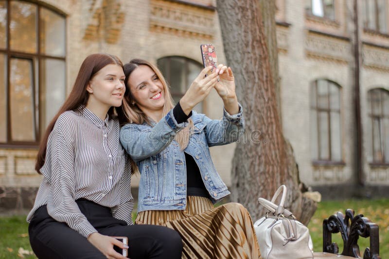 Zwei schöne junge Mädchen, die selfie am Telefon beim Sitzen auf einer Bank im Herbstpark nahe der Universität nehmen lizenzfreies stockfoto Zwei schöne junge Mädchen, die selfie am Telefon beim Sitzen auf einer Bank im Herbstpark nahe der Universität nehmen lizenzfreies stockfoto