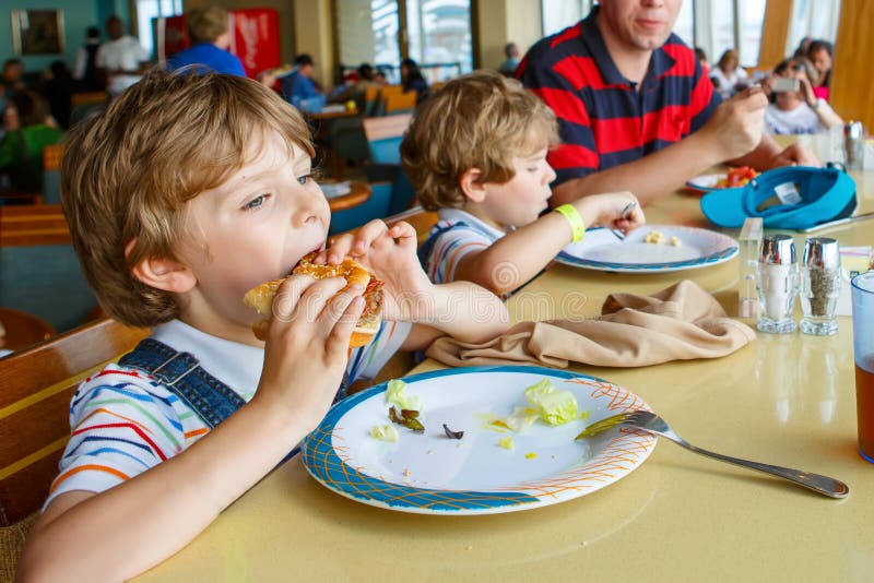 Zwei Vorschulkinder und Vaterjungen, die den Teigwarenhamburger sitzt im Café auf Kreuzschiff essen Glückliche Kinder, Zwillinge