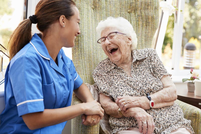 Ältere Frau, die im Stuhl sitzt und mit Krankenschwester In Retirement Home lacht