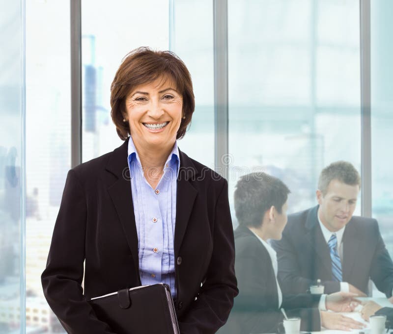 Senior businesswomanstanding in office holding personal organizer. Businesspeople working at desk in background. Senior businesswomanstanding in office holding personal organizer. Businesspeople working at desk in background.