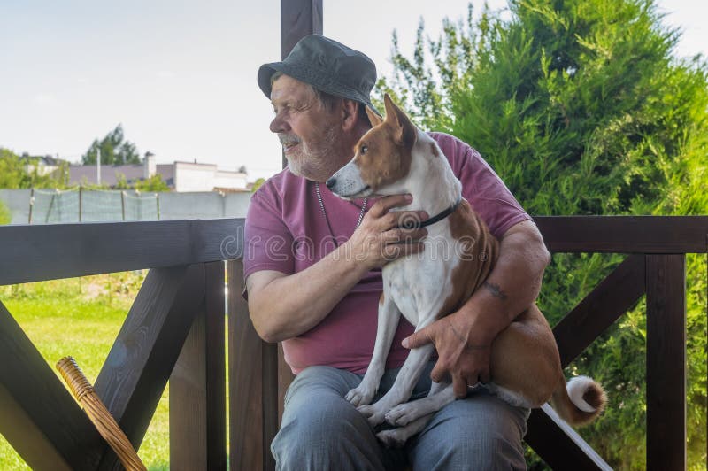 Älterer Mann, der seinen reizenden basenji Hund nimmt und beide beim Sitzen auf Sommerveranda aufpasst lizenzfreie stockbilder Älterer Mann, der seinen reizenden basenji Hund nimmt und beide beim Sitzen auf Sommerveranda aufpasst lizenzfreie stockbilder