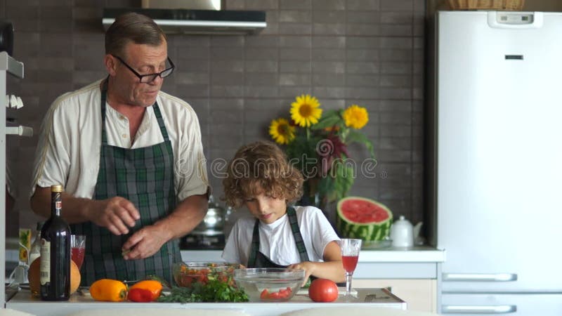Älterer Mann gibt dem Jungen ein Küchenmesser und bringt Ihnen bei, wie man eine Gurke schneidet Großvater und Enkel in der Küche