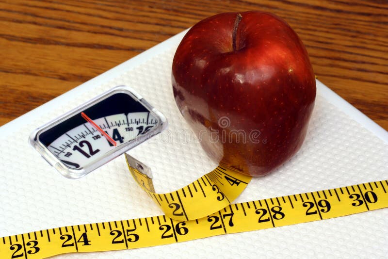 A tape measure wraps around a white bathroom scale which has an apple on top. A tape measure wraps around a white bathroom scale which has an apple on top.