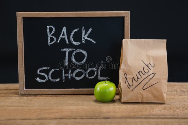 Lunch paper bag, green apple and slate with text back to school on wooden table against black background. Lunch paper bag, green apple and slate with text back to school on wooden table against black background