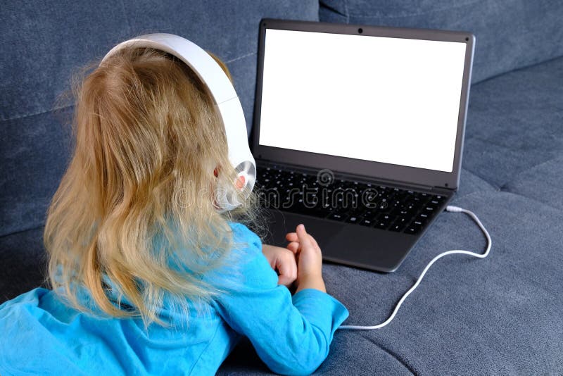 Child 2-3 years old, girl in headphones lies on sofa in front of laptop, uses Internet, watches educational cartoons, plays computer games, white blank screen for video, concept of remote learning. Child 2-3 years old, girl in headphones lies on sofa in front of laptop, uses Internet, watches educational cartoons, plays computer games, white blank screen for video, concept of remote learning