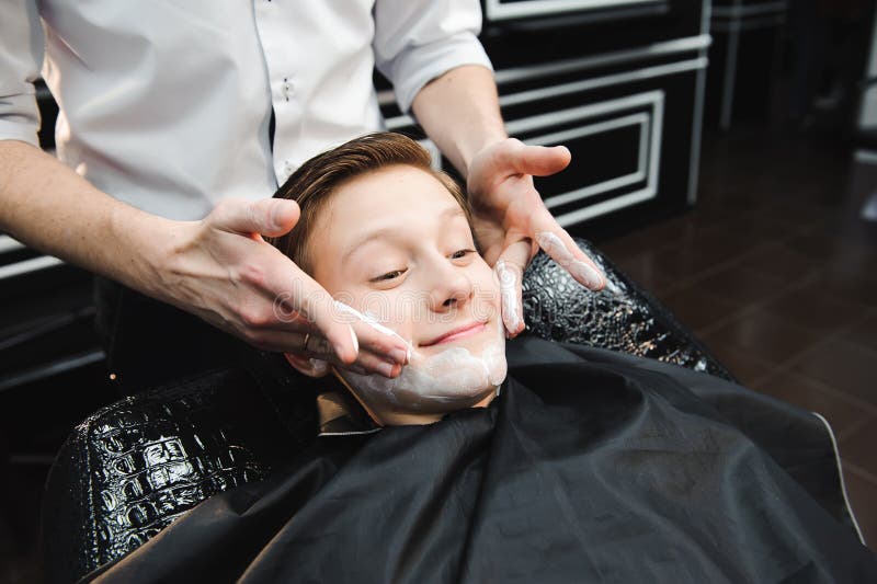 Śmieszna chłopiec w czarnym salonu przylądku w zakładzie fryzjerskim Fryzjer męski stosuje golenie pianę z pomocą golenia muśnięc zdjęcia stock Śmieszna chłopiec w czarnym salonu przylądku w zakładzie fryzjerskim Fryzjer męski stosuje golenie pianę z pomocą golenia muśnięc zdjęcia stock