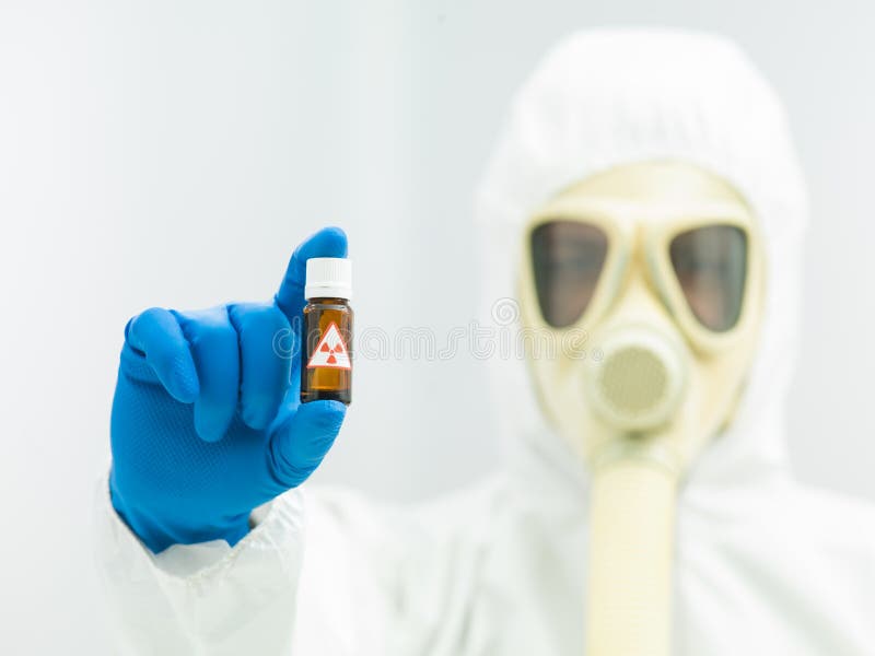 Radioactive isotope sample in a small brown bottle with a warning label held in the fingers of a laboratory scientist in full protective clothing with a mask and breathing apparatus. Radioactive isotope sample in a small brown bottle with a warning label held in the fingers of a laboratory scientist in full protective clothing with a mask and breathing apparatus