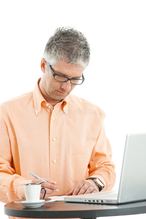 Casual businessman wearing orange shirt and jeans, standing at coffee table, using laptop computer, writing notes. Isolated on white. Casual businessman wearing orange shirt and jeans, standing at coffee table, using laptop computer, writing notes. Isolated on white.