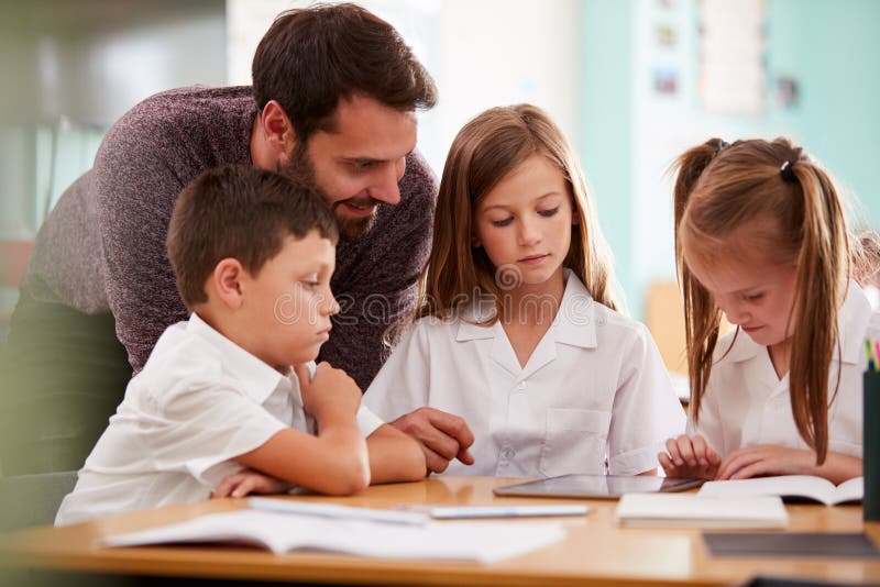 Élèves d'école de With Three Elementary de professeur masculin portant l'uniforme utilisant la Tablette de Digital au bureau images libres de droits Élèves d'école de With Three Elementary de professeur masculin portant l'uniforme utilisant la Tablette de Digital au bureau images libres de droits