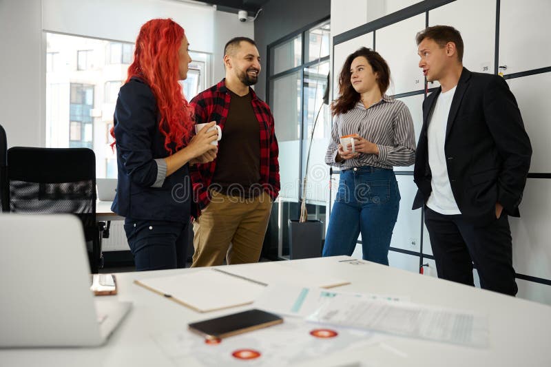 Friendly team of marketers resting and laughing, having talks during coffee break, gathering near lockers in coworking office, young business company. Friendly team of marketers resting and laughing, having talks during coffee break, gathering near lockers in coworking office, young business company