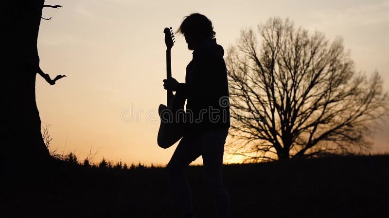 Équipez la guitare électrique de jeux et chantez une chanson lyrique dans un domaine près de l'arbre au coucher du soleil Silhoue