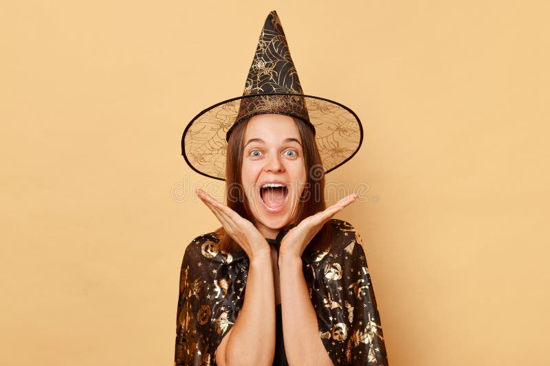 Amazed excited young woman wearing witch costume and carnival cone hat celebrating halloween isolated over beige background keeps hand under chin screaming with excitement. Amazed excited young woman wearing witch costume and carnival cone hat celebrating halloween isolated over beige background keeps hand under chin screaming with excitement