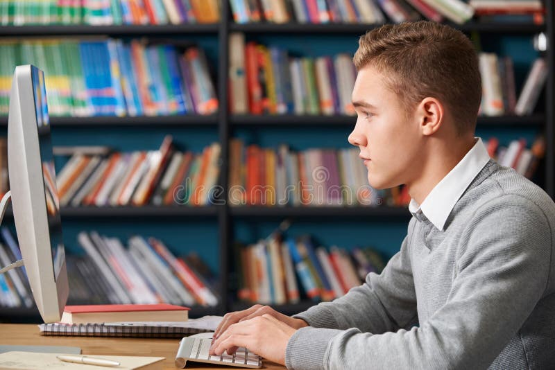 Étudiant adolescent masculin Working At Computer dans la bibliothèque photo libre de droits Étudiant adolescent masculin Working At Computer dans la bibliothèque photo libre de droits