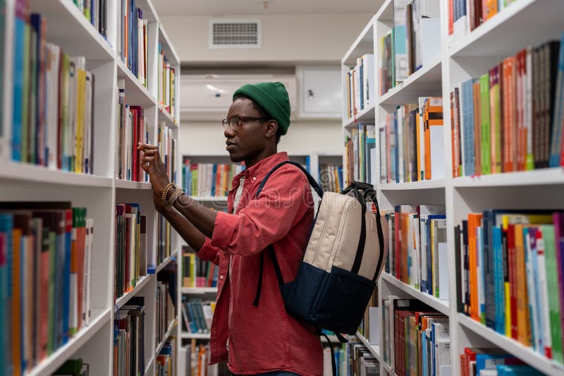 étudiant africain réfléchi homme en lunettes choisir livre dans la bibliothèque de collège ou librairie photos libres de droits