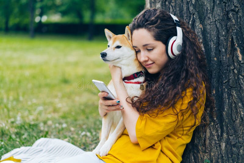 Étudiant de sourire appréciant la musique dans des écouteurs utilisant le smartphone en parc avec le chien images libres de droits Étudiant de sourire appréciant la musique dans des écouteurs utilisant le smartphone en parc avec le chien images libres de droits
