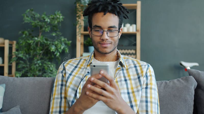 Étudiant heureux afro-américaine en utilisant le téléphone intelligent à la maison reposant sur le canapé banque de vidéos