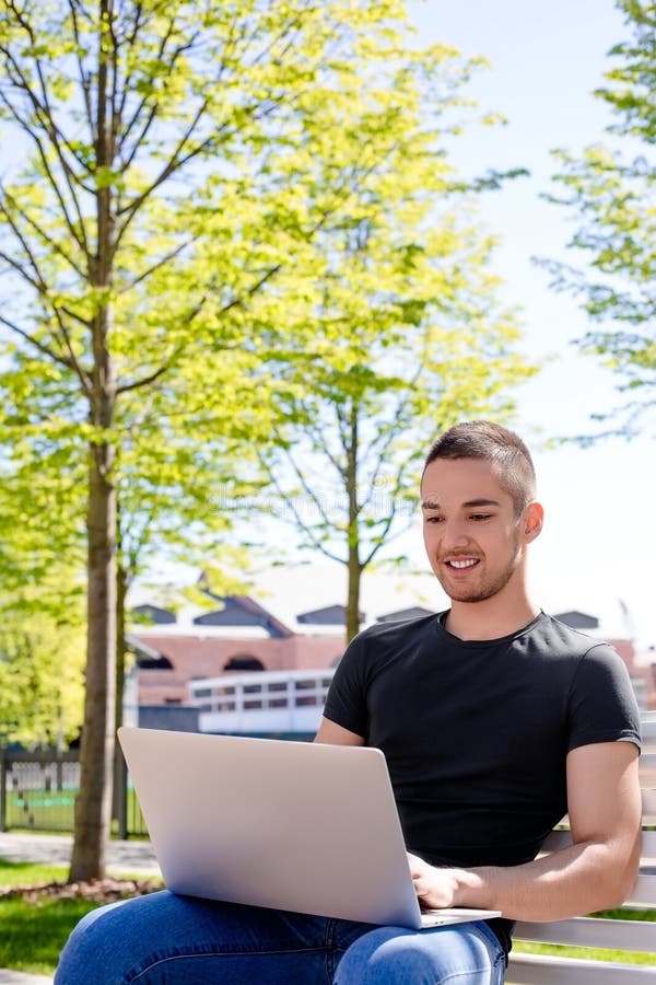 Étudiant masculin joyeux ayant l'éducation en ligne par l'intermédiaire du netbook, se reposant sur un campus photo libre de droits Étudiant masculin joyeux ayant l'éducation en ligne par l'intermédiaire du netbook, se reposant sur un campus photo libre de droits
