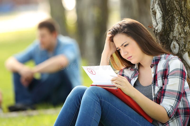 Single sad student looking at failed exam sitting on the grass in a park with unfocused people in the background. Single sad student looking at failed exam sitting on the grass in a park with unfocused people in the background