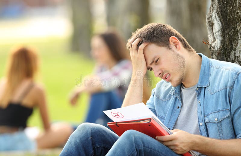 Single sad student checking a failed exam sitting on the grass in a park with unfocused people in the background. Single sad student checking a failed exam sitting on the grass in a park with unfocused people in the background