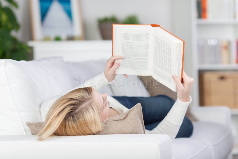 Étudiante Reading Book While se trouvant sur le sofa photographie stock libre de droits Étudiante Reading Book While se trouvant sur le sofa photographie stock libre de droits