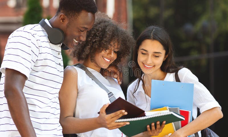 Cheerful university friends studying together outdoors, standing with workbooks in college campus, checking classes schedule. Cheerful university friends studying together outdoors, standing with workbooks in college campus, checking classes schedule