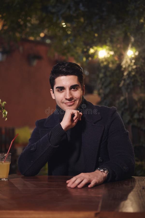 Één jong knap mensenportret, die in openlucht in koffietuin bij lijst, nacht zitten die, aan camera kijken Hoger lichaamsschot stock foto's Één jong knap mensenportret, die in openlucht in koffietuin bij lijst, nacht zitten die, aan camera kijken Hoger lichaamsschot stock foto's