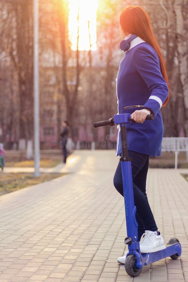 ??n jonge Kaukasische vrouw met rood haar in een blauwe laag op een blauwe elektrische autoped in het park Milieuvriendelijk verv royalty-vrije stock foto ??n jonge Kaukasische vrouw met rood haar in een blauwe laag op een blauwe elektrische autoped in het park Milieuvriendelijk verv royalty-vrije stock foto